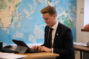 Senior boy sitting in a classroom working on a digital tablet with a map on the wall